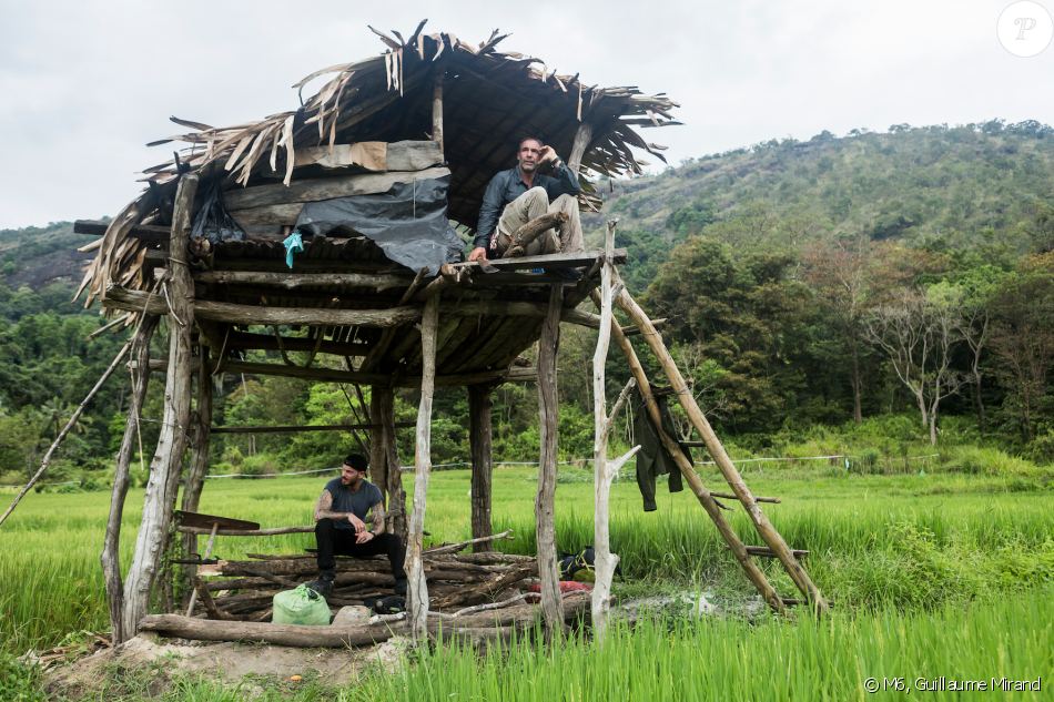 Two men relax on a wooden structure in a lush green rice field, surrounded by mountains and trees under a cloudy sky.
