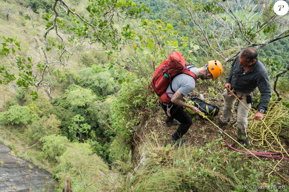 Two climbers prepare ropes and gear on a rocky ledge surrounded by lush green foliage